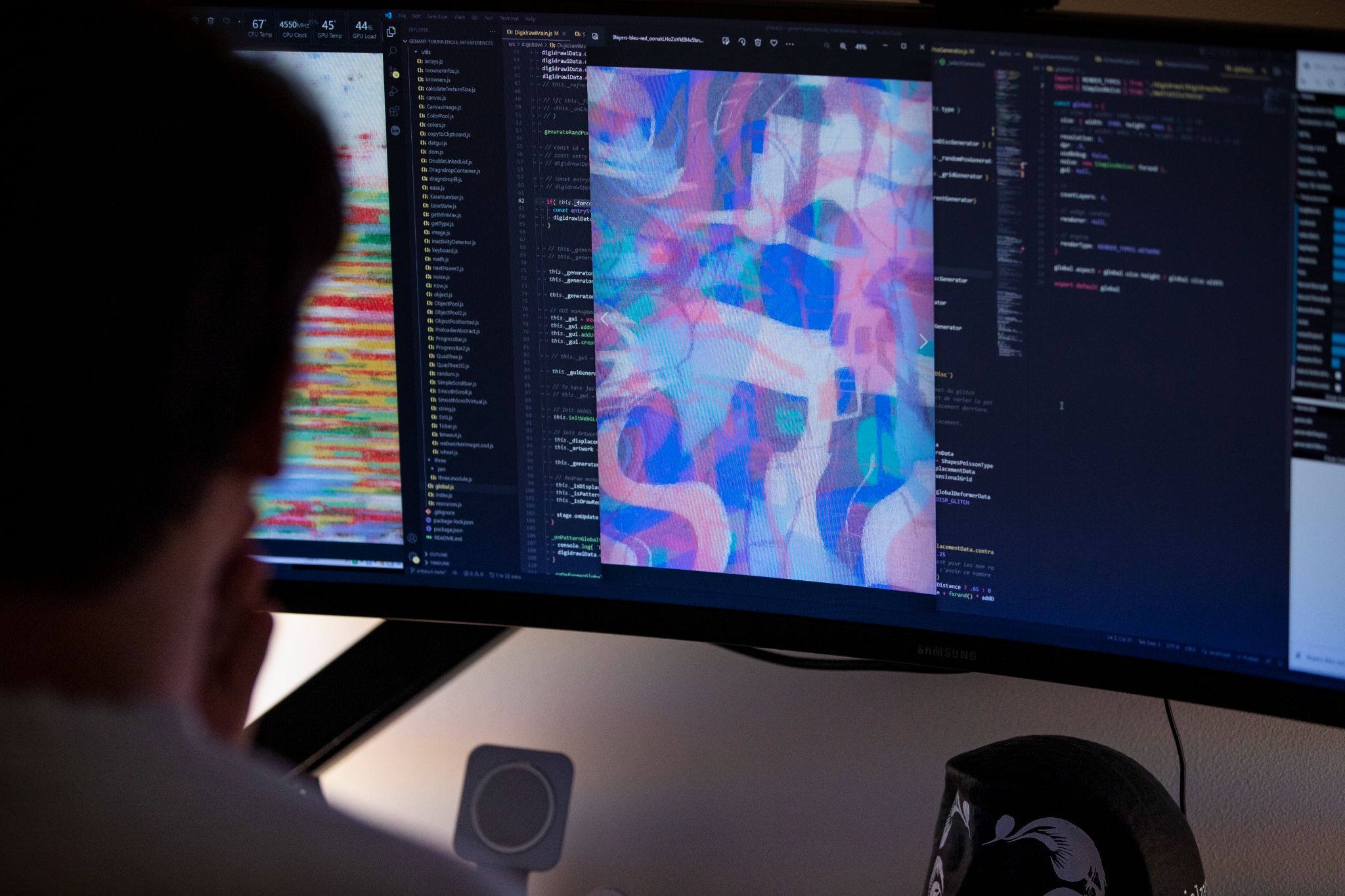 Artist working with code and generative artwork displayed on a wide monitor — code on left, vibrant abstract output in center